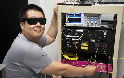 Yicheng Shi wears dark safety glasses as he poses next to a rack of electronics equipment.