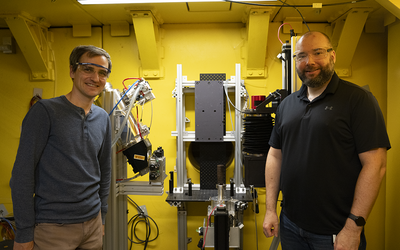 Cyrus Daugherty and Jake LaManna, wearing safety glasses, pose on either side of an imaging device holding a replica scroll.
