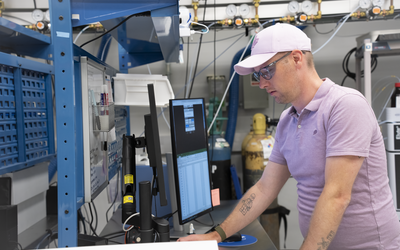 A man wearing safety glasses and a baseball cap leans over a lab table holding computer monitors that show spreadsheets of data.