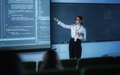 A female instructor wearing glasses presents code on a large screen to a classroom.