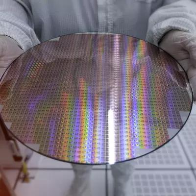 A person in coveralls and white gloves holds out a circular wafer of reflective silicon chips. 