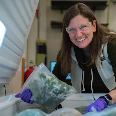 Melissa Phillips, wearing safety glasses, poses leaning over an open chest freezer, holding a plastic bag of spinach. 