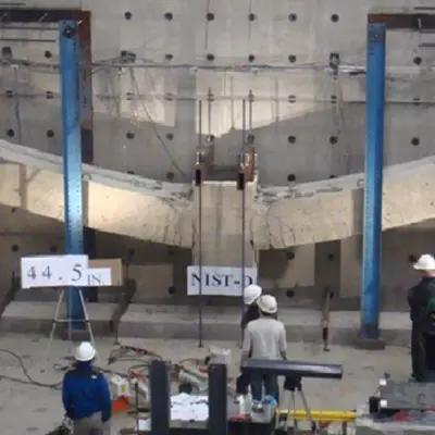 Researchers in hard hats stand inside a large indoor space studying a horizontal concrete test structure on a wall.