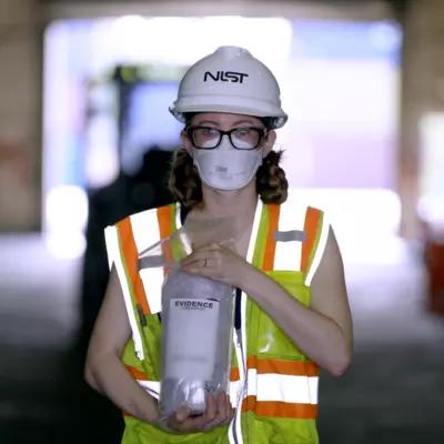 A woman in a NIST hard hat and other safety gear stands inside a warehouse holding a plastic bag labeled "Evidence."