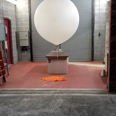 A large white balloon with a small box attached to it waits inside a cinderblock building with a rolltop door. 