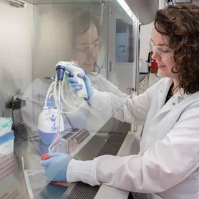 Jerilyn Izac sits at a lab bench with her hands inside a plastic shield, using a device to draw liquid from a plastic container into a tube. 