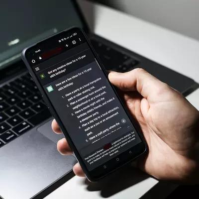 In front of a laptop computer, a hand holds a cell phone that has a conversation with generative AI on the phone screen.