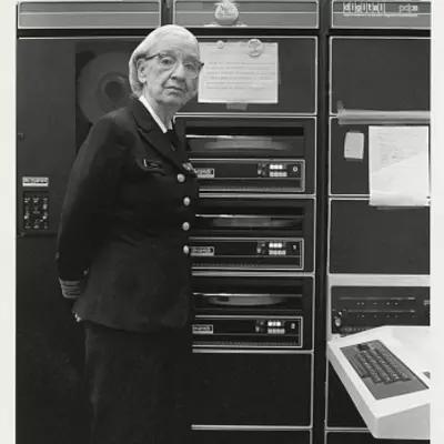 Historical photo shows Grace Hopper in uniform standing next to a rack of computer equipment.