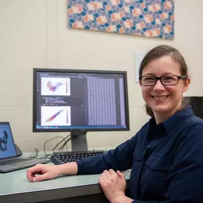 Melinda Kleczynski poses sitting at her desk, with a monitor showing data and graphs behind her. 