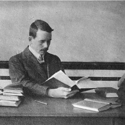 Historical photo shows Henry Moseley sitting at a desk reading a thick book, with other open books on the desk. 