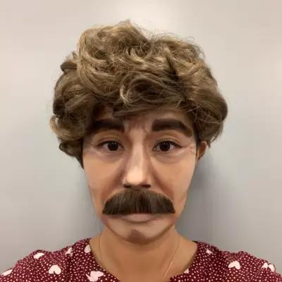 A woman is disguised as a man with contoured makeup, a moustache and a wig.