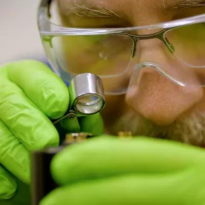 A researcher wearing green gloves and safety glasses over his regular glasses holds a small magnifying glass close to his face to look at a hard drive.