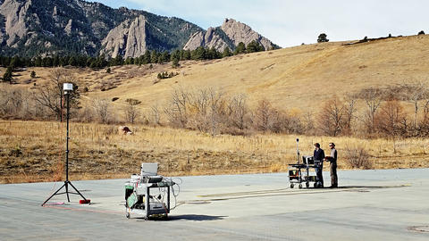 Mountains in the background. Left: rolling cart with equipment. Tall tripod. Right: Two men with another rolling cart of equipment.