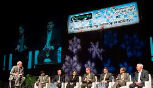 Moderator sits on the left. Seven people (6 men, 1 woman) sit in chairs.