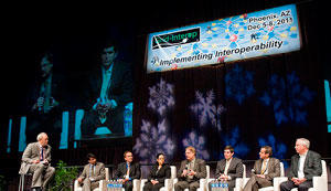 Moderator sits on the left. Seven people (6 men, 1 woman) sit in chairs.