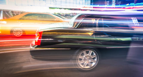 A streaked image of a taxi and a dark car driving in Times Square, New York City street at night.