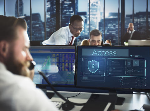 man on a phone sitting in front of  two computer screens. Other men on computers in the background