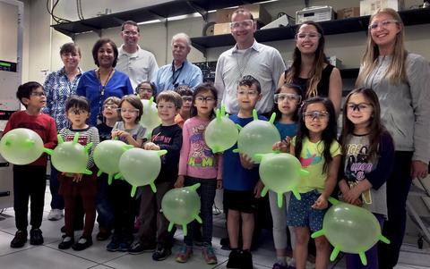 Adults standing in the back row. Kids with blown up green gloves stand in the front row.