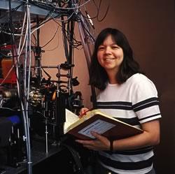 Physicist Deborah Jin in front of equipment holding an open lab  notebook.