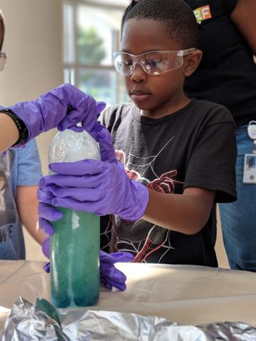 Young boy wearing purple gloves and safety goggles holds a plastic bottle filled with aqua colored liquid as another hand screws on the cap.