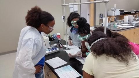 Jeanita and four young girls, all in safety glasses, stand around a lab table, their notebooks open. 