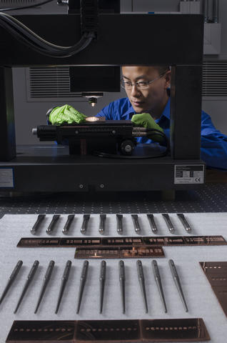 A man with green gloves looks at an item under a microscope