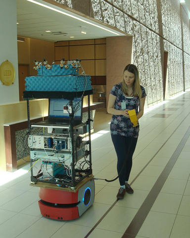 Woman in large indoor hallway standing next to tall stack of electronic equipment.
