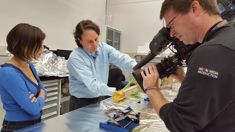 Scientist doing work while to his left is a reporter and in front of him is a camera man.