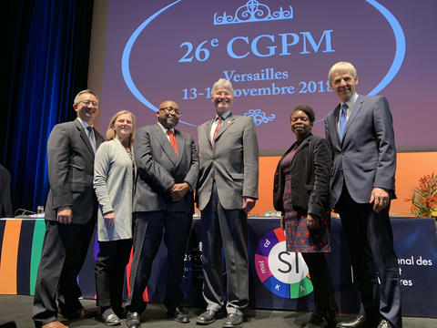 6 people standing on a stage in front of a screen that reads: 26th CGPM