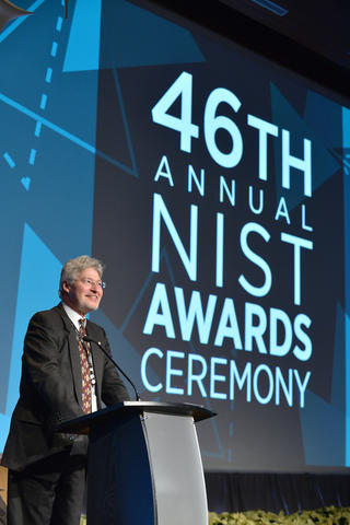 A man stands at a podium in front of a large audience, and behind him a screen is lit up with the words 46th Annual NIST Awards Ceremony."