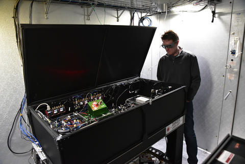 Man in goggles standing next to and looking into a large box with open lid showing what appears to be electrical equipment and wiring inside