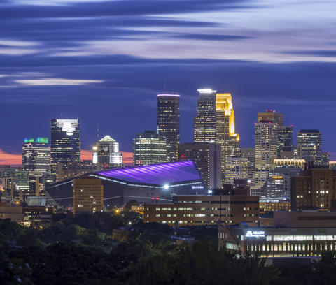 The darkened skyline of a city, with a stadium in the middle. The stadium glows purple.