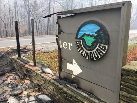 The Great Smoky Mountains National Park Gatlinburg Welcome Center sign is half-burned, leaving only the Gatlinburg name and a white arrow. Debris lies on the ground in front of the sign.