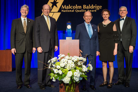 Center: Baldrige award on a podium. Left: 2 men in suits. Right: a man in a suit, woman in dress, man in suit