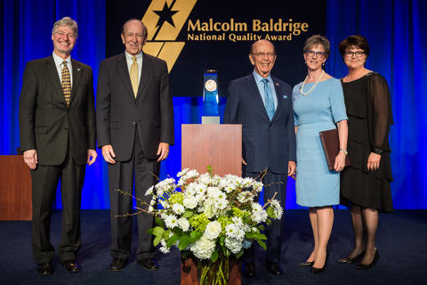 Center: Baldrige Award on a podium. Left: two men in suits. Right: a man in a suit and two women in dresses