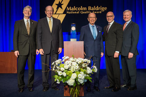 Center: Baldrige Award on a podium. Left: 2 men in suits. Right: 3 men in suites