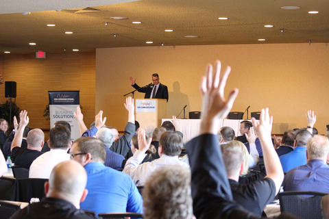 Man stands at a podium. Backs of people with raised hands