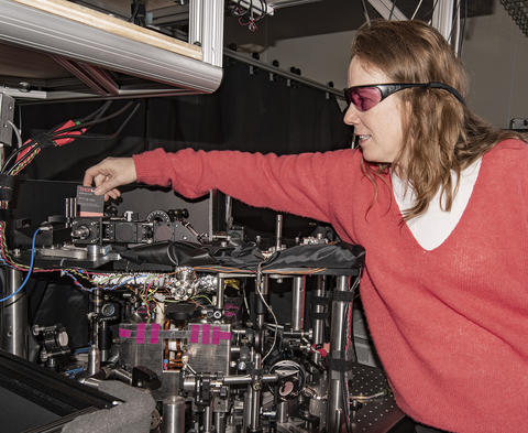 Woman with reddish hair and wearing a pink sweater and laser safety glasses reaching over an instrument table and holding a card used to path of lasers.