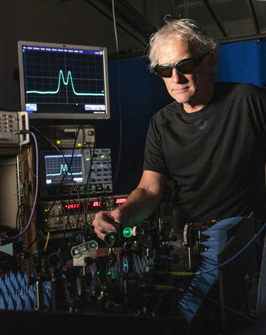 Man in black shirt and dark glasses facing camera is adjusting a mirror on a table of lasers and optics. On the left behind him are several electronics boxes topped by a computer monitor showing a blue signal that looks like cats ears.
