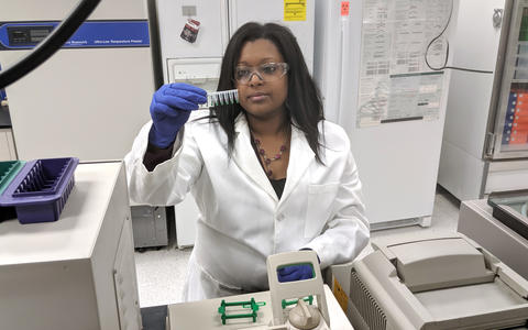 woman in a lab coat looks at vials with a small amount of green liquid in them before putting them into a machine