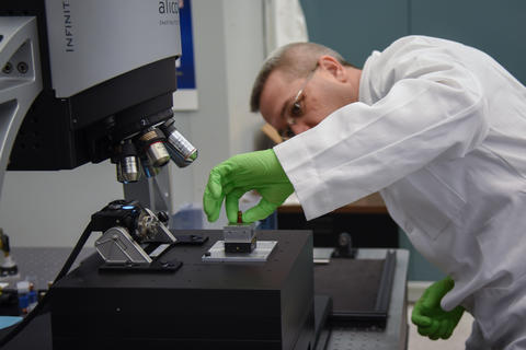 A man in a lab coat and green gloves places a bullet under a microscope.