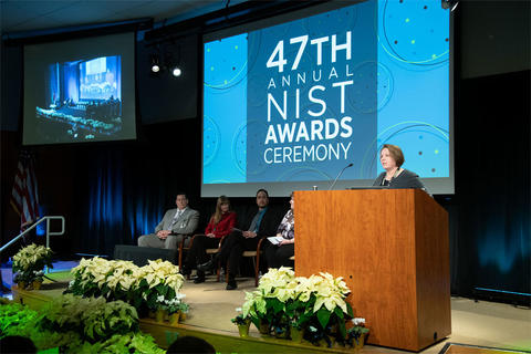 A woman stands behind a podium onstage with screen behind her saying: 47th Annual NIST Awards.