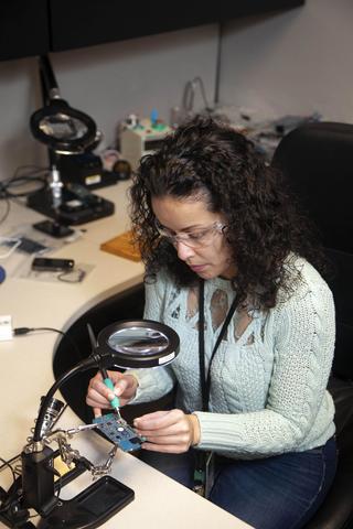 A woman uses a soldering iron to attach wires to a circuit board.