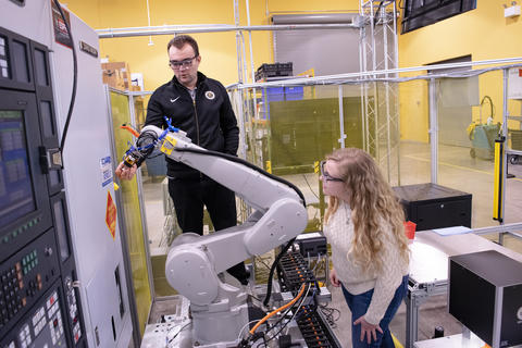 A man stands behind a robot arm and a woman stands next to it. Both are watching what it's doing.