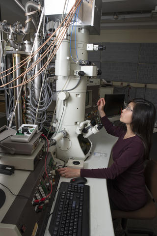 June Lau with retrofitted microscope NIST researcher in a laboratory room looking at tall vertical microscope with multicolored wires emanating from it.