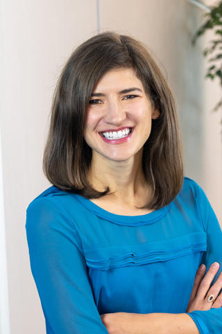 A headshot of Sarah Hughes, a woman smiling with arms crossed, wearing a blue shirt