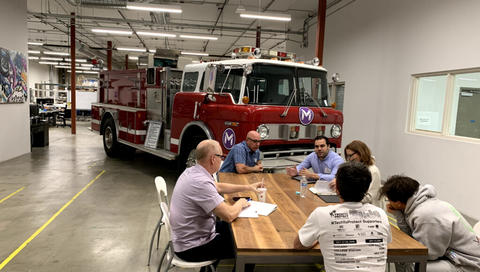 This image shows a group of people sitting around a wooden table with a firetruck behind them