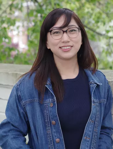 Portrait of Grace Hahm, a young woman with medium length dark hair and glasses, wearing a jean jacket and smiling for a photo outside.