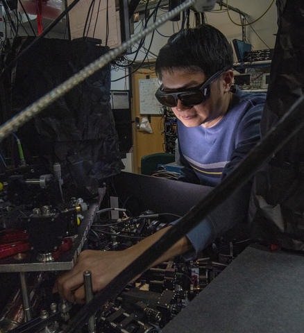 Man with dark hair and wearing a blue shirt and laser safety goggles standing on the right and facing to the left as he reaches into a two-level laboratory table full of optics and other equipment. 