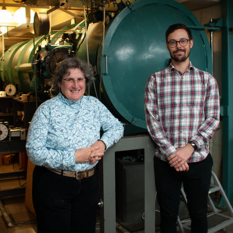 A woman and a man stand in front of scientific equipment, including a large green tank. 